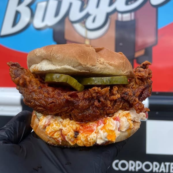 A Rockin' Burgers fried chicken sandwich with pickles and creamy slaw, held by a gloved hand.