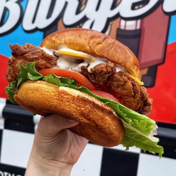 Hand holding a Rockin' Burgers fried chicken sandwich with lettuce, tomato, cheese, and sauce.