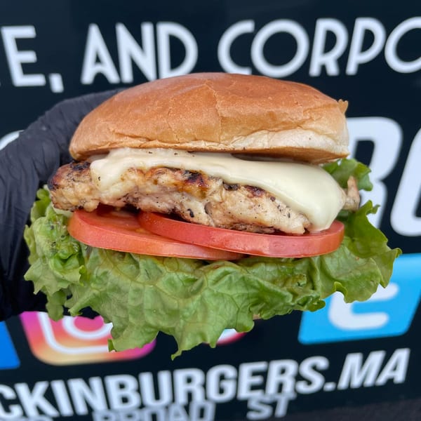 A hand in a black glove holds a Rockin' Burgers chicken sandwich with melted cheese, lettuce, and tomato.